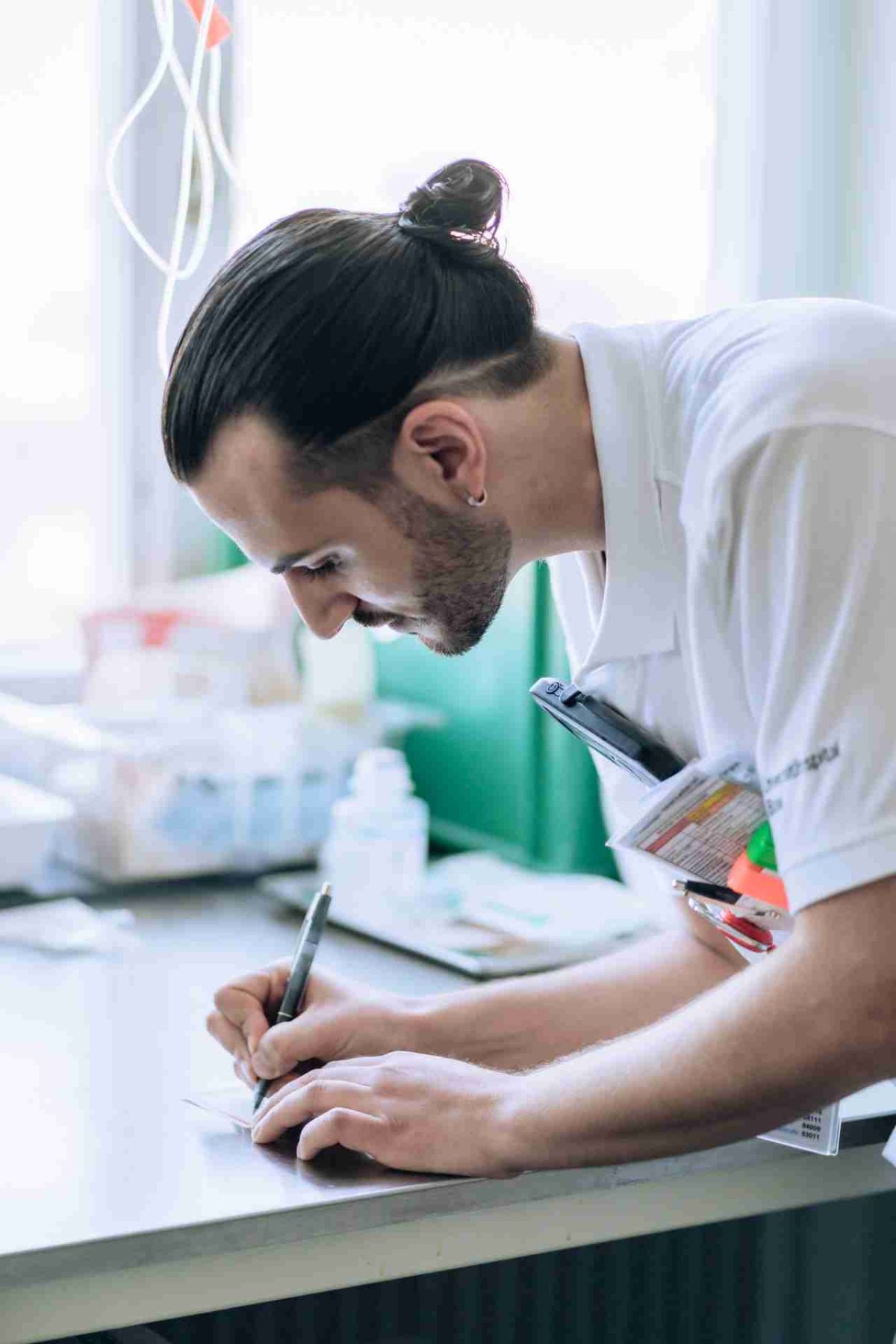 Ein Mann mit zurückgebundenem Haar und Ohrring, gekleidet in ein weisses Hemd, steht an einem Tisch und schreibt konzentriert mit einem Stift auf Papier. In seiner Hemdtasche sind Stifte und Dokumente verstaut. Der Hintergrund zeigt eine helle, leicht unscharfe Arbeitsumgebung mit medizinischen Utensilien.