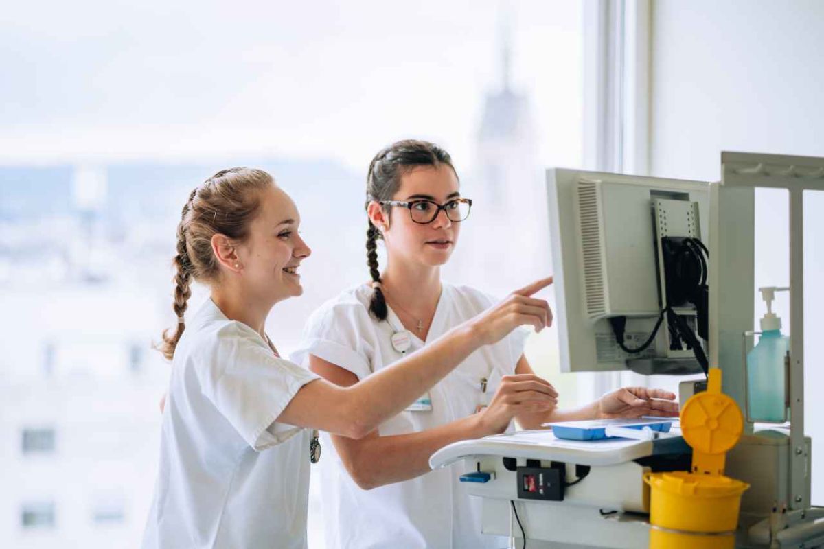 Zwei junge Frauen in weißer Berufskleidung stehen vor einem Computerbildschirm und arbeiten gemeinsam daran. Eine von ihnen zeigt auf den Bildschirm, während die andere lächelt. Beide tragen geflochtene Zöpfe und haben Namensschilder. Im Hintergrund ist ein helles Fenster mit einer verschwommenen Aussicht auf eine Stadtlandschaft zu sehen. Auf dem Tisch befinden sich medizinische Geräte und Utensilien, darunter ein Desinfektionsspender. Die Szene vermittelt eine kooperative und professionelle Arbeitsatmosphäre.